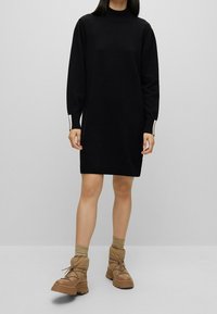Robe noire en maille avec un col montant, des manches longues et des poignets côtelés. Portée avec des bottes matelassées couleur sable dotées d'une semelle épaisse et de lacets.