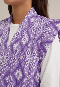 Lila mönstrad sjal med vita geometriska mönster, i ett texturerat tyg och breda slag, lagd över en vanlig vit topp.