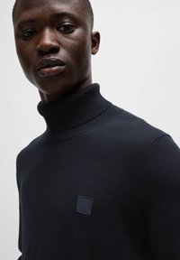 Pleteni pulover s turtleneck ovratnikom v temno mornarsko modri barvi, z rebrastim ovratnikom, majhnim kvadratnim logotipom na prsih, mehko teksturo in prilegajočo zasnovo.
