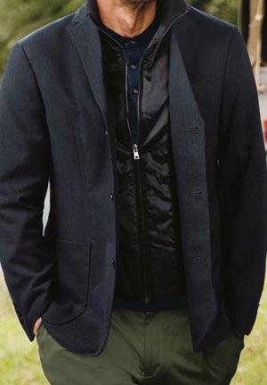 Marineblazer mit taillierter Passform, ausgestattet mit zwei Vorderaschen, über einer schwarzen Zip-Jacke getragen. Glatte Stoffstruktur, modernes Design.
