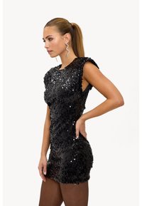 Zwart mini-jurkje met pailletten, cap sleeves, nauwsluitend ontwerp en een gestructureerd oppervlak. Model draagt een slangvormige oorbel en donkere panty's.