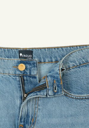 Gros plan sur un jean en denim bleu clair avec la braguette partiellement dézippée, un bouton en bois, et une étiquette en tissu noir portant l'inscription « FAGUO DENIM ».