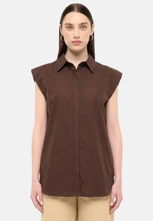 Femme aux cheveux longs et foncés portant une chemise marron sans manches à col et un pantalon beige, debout devant un fond clair uni.