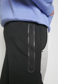 Svart och grå joggers i en tvåfärgad design, med en sidofick med dragkedja och svarta detaljer. Materialet verkar mjukt med en matt textur.