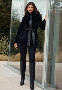 Melnas mākslīgās ādas jaka ar apkakli un jostu; apvienota ar melnām legingām un augstiem papēžiem līdz ceļgaliem. Gluda tekstūra un eleganta forma.