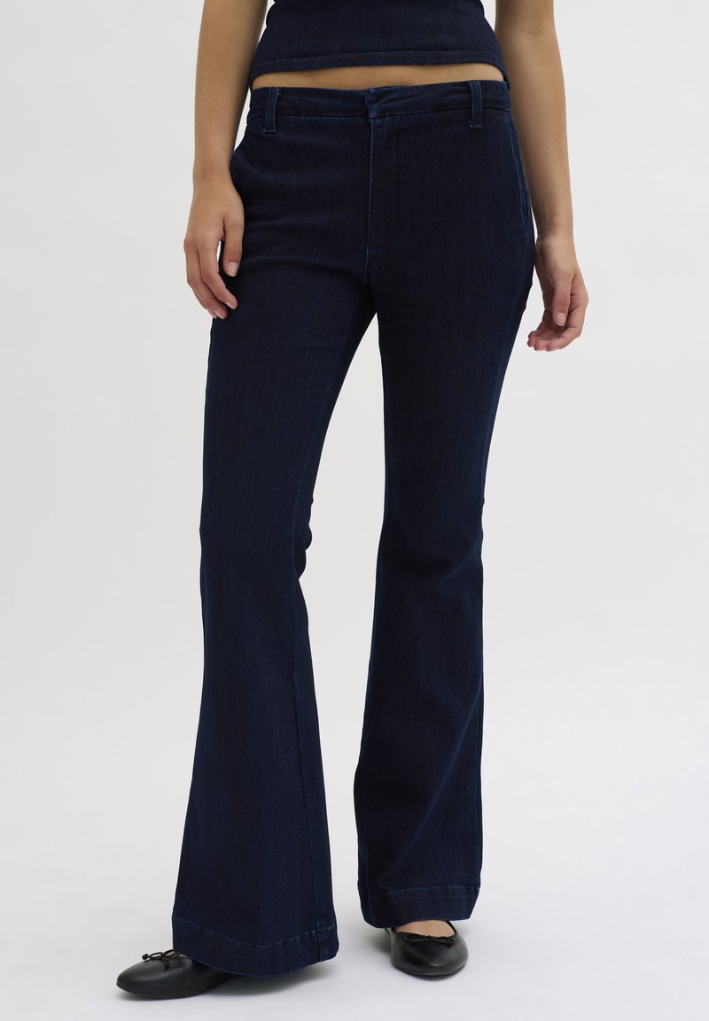 Dark denim flared pants with a fitted waist, front pockets, and clean stitching. Fabric has a smooth texture and subtle vertical lines.