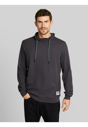Man met kort krullend haar, gekleed in een donkergrijze hoodie met aantrekkoorden, met één hand in de zak, staand tegen een grijze achtergrond.