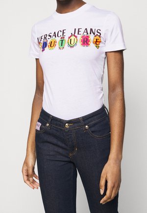 Person, die ein weißes Versace Jeans Couture T-Shirt mit bunten Logo-Buchstaben und dunkelblauen Jeans trägt, vor einem schlichten Hintergrund stehend.