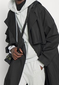 Abrigo trinchera negro recortado sobre una sudadera con capucha gris claro y pantalones, con herrajes en contraste y un ajuste relajado.