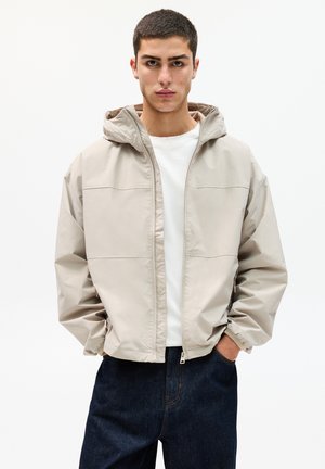 Beige huva jacka med dragkedja framtill, med elastiska ärmslut och en avslappnad passform. Sammanförd med mörka jeansbyxor. Slät textur.