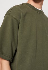 Sudadera de algodón verde oliva con cuello redondo, mangas cortas y puños de canalé. Textura suave con un ajuste relajado. Sin patrones visibles.