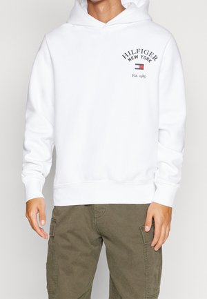 Mężczyzna w białej bluzie z kapturem Hilfiger New York z logo i napisem na piersi, w połączeniu z oliwkowymi spodniami typu cargo, stoi nieruchomo.