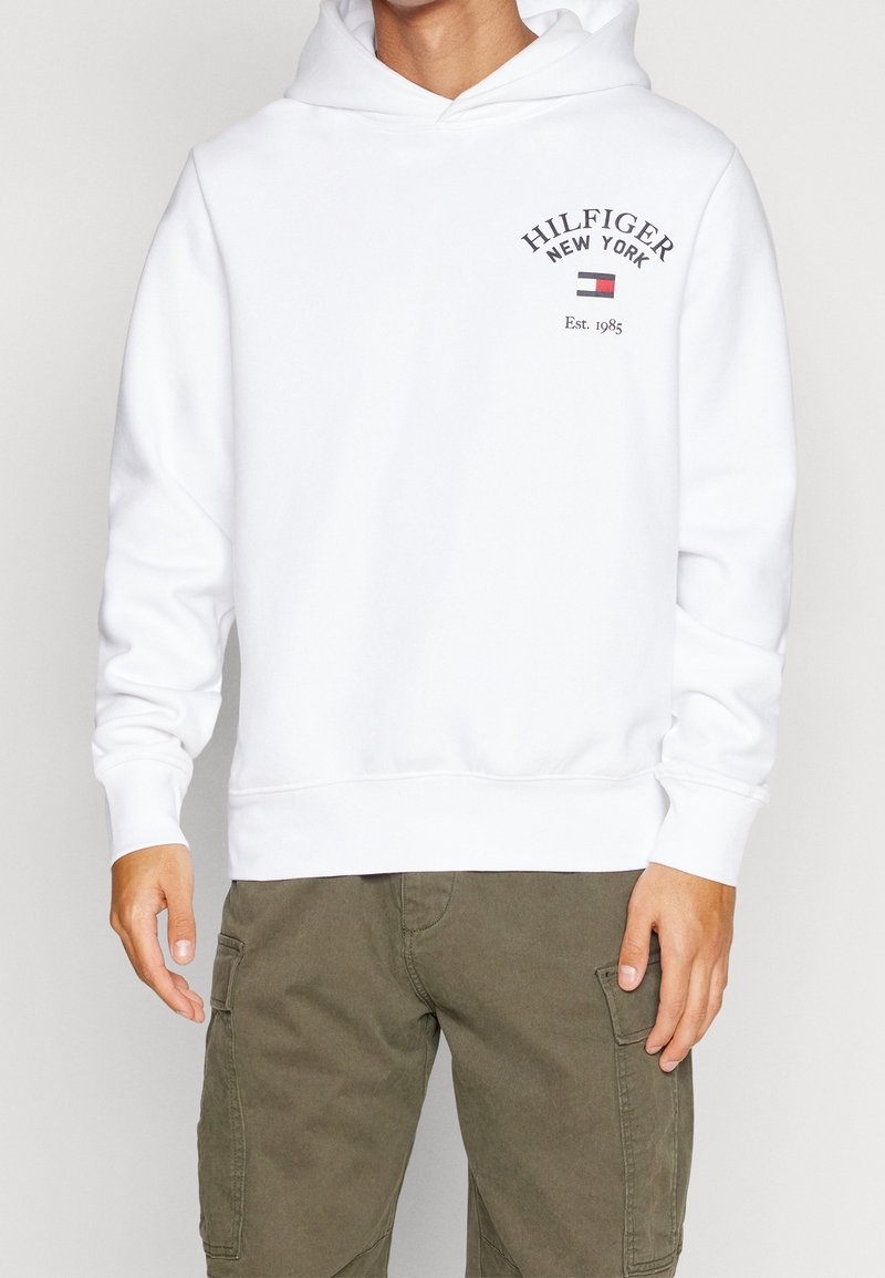 Homme portant un hoodie blanc Hilfiger New York avec un logo et un texte sur la poitrine, associé à un pantalon cargo vert olive, debout immobile.