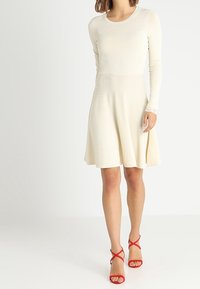 Knee-length, krämfärgad klänning i ett mjukt stickat material med långa ärmar och en åtsittande bodice som vidgar sig till en cirkulär kjol. Röda sandaler med remmar.