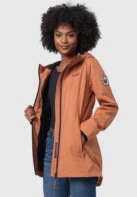 Giacca arancione impermeabile con cappuccio, zip frontale e rifiniture nere; presenta dettagli con logo e toppe, tessuto leggero e tasche laterali.