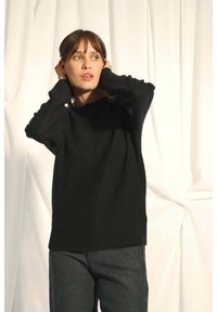 Musta, neulottu villapaita, jossa on leveä kaula-aukko ja pitkät hihat, yhdistettynä tummiin farkkuihin. Pehmeä rakenne, minimalistinen muotoilu, ei näkyviä kuvioita.