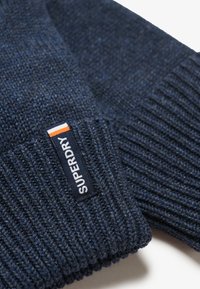 Gorro de punto en color azul marino con textura acanalada. Presenta una etiqueta negra elevada con detalles en blanco y naranja, de la marca "SUPERDRY".