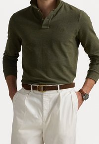 Olive green long-sleeve polo shirt with a collar, two-button placket, and subtle texture, paired with white trousers and a brown belt.