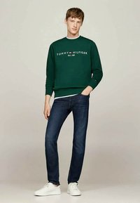 Joven vestido con un sudadera oscura verde de Tommy Hilfiger, jeans oscuros azules y zapatillas blancas, de pie con una mano en el bolsillo.