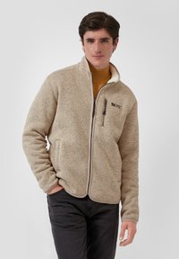 Junger Mann in einer beigen Fleecejacke mit Reißverschluss über einem senfgelben Rollkragenpullover, mit einer Hand in der Tasche, stehend vor einem weißen Hintergrund.