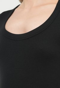 Κοντινό πλάνο ενός ατόμου που φοράει μαύρο ριπαριστό τοπ με φαρδιά λαιμόκοψη σε σχήμα scoop, που δείχνει το άνω μέρος του στήθους και της περιοχής του λαιμού.