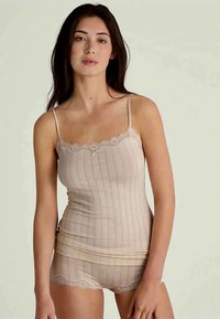 Ung kvinne iført en beige ribbet cami-top og matchende shorts med blondekant, stående mot en ensfarget lys bakgrunn.