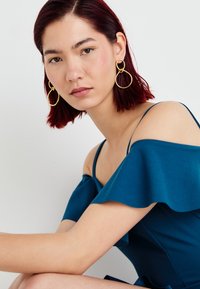 Vestido teal sem alças com mangas com volantes, textura suave. A modelo usa grandes brincos de ouro em forma circular, cabelo liso e maquiagem neutra.
