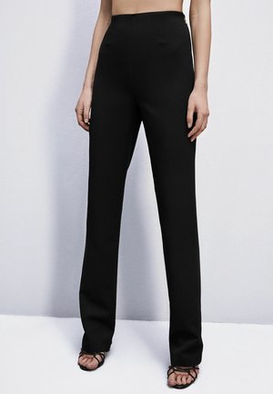 Kobieta ubrana w czarne, proste spodnie z wysokim stanem i czarne sandały na szpilce, stojąca na tle gładkiej, jasnej ściany.