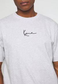 Hombre con camiseta gris claro con pequeño texto en cursiva negro "Kani" centrado en el pecho, contra un fondo liso.