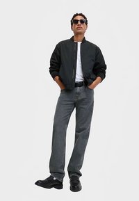 Černá bomber bunda přes bílé tričko, doplněná volnými šedými džínami a černými botami, doplněná tmavými slunečními brýlemi.