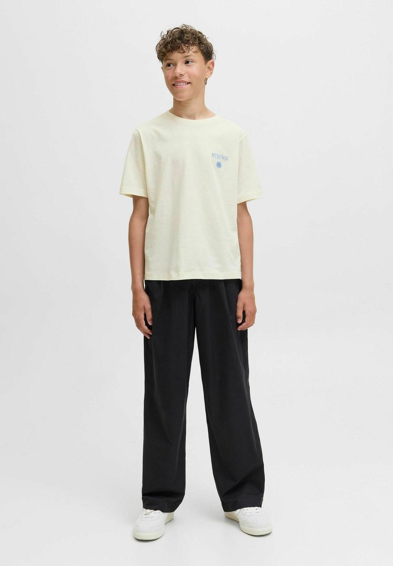 Camiseta de manga corta amarilla claro con texto azul en el lado izquierdo del pecho, combinada con pantalones anchos negros y zapatillas blancas. Diseño simple.