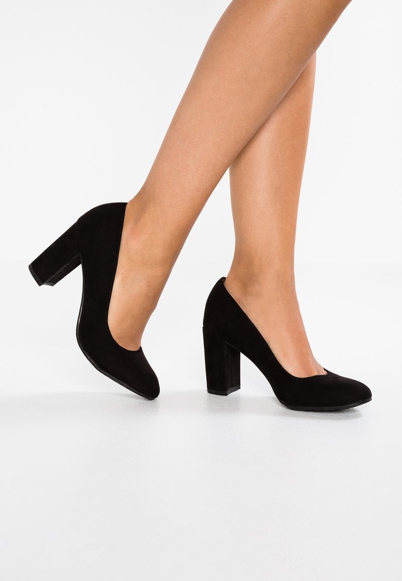 Jambes chaussées de chaussures en daim noir à talons blocs, un pied à plat et l'autre soulevé sur les orteils sur fond blanc.