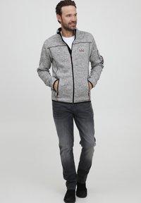 Grå zip-up jakke med sorte accenter, fremstillet af et tekstureret stof, med to sidelommer og en stående krave. Båret med mørke jeans og sorte sko.