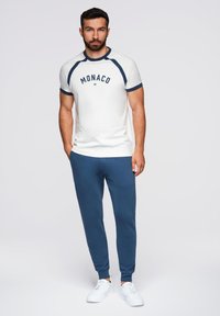 Witte T-shirt met blauwe accenten en de tekst "MONACO", gecombineerd met blauwe joggers. Heeft een sportieve pasvorm en een ronde hals. Witte sneakers.