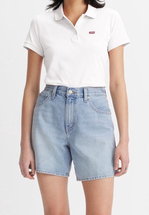 Frau trägt ein weißes Poloshirt mit kurzen Ärmeln und hellblaue Jeans-Shorts mit hohem Bund und Vordertaschen.