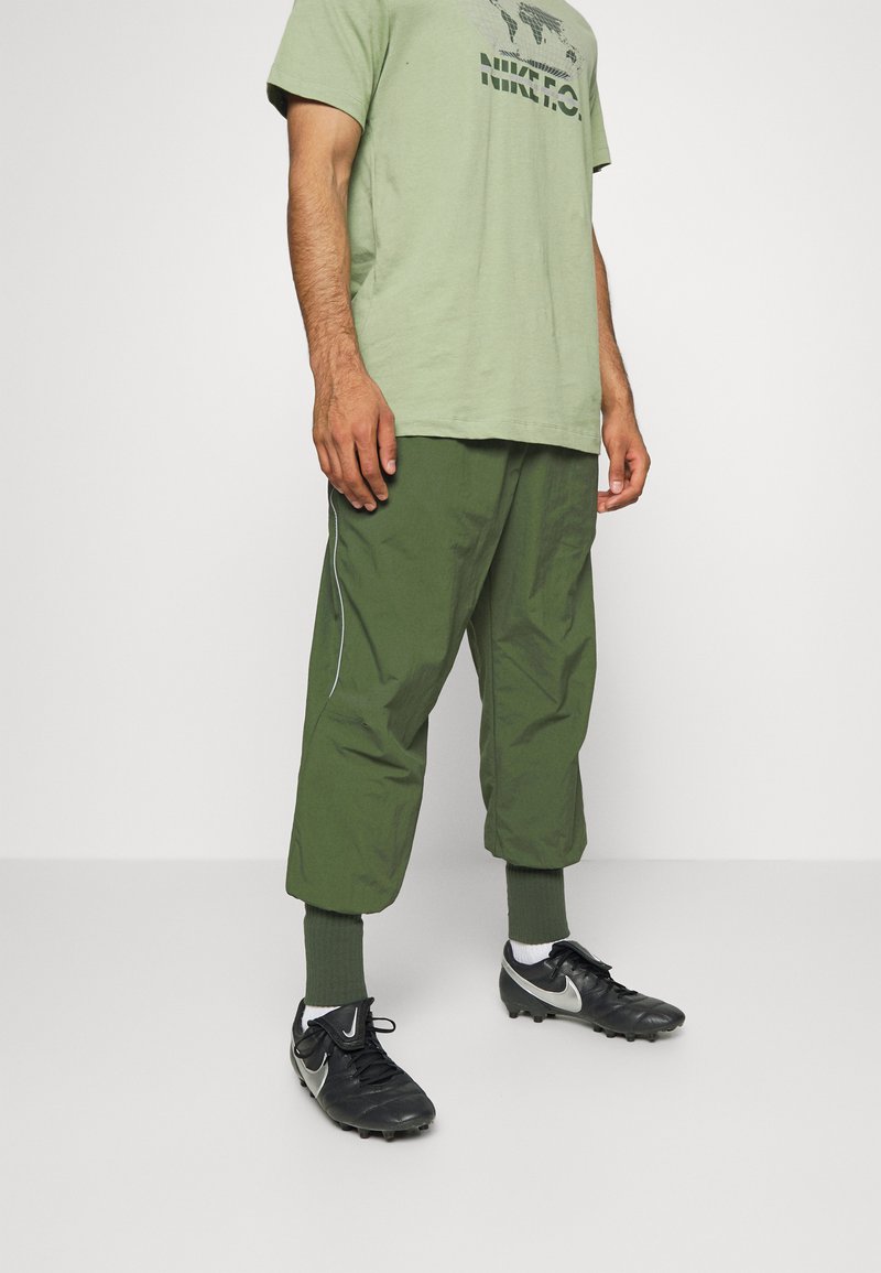 Grünes Baumwoll-T-Shirt mit schwarzfarbenem Logo, kombiniert mit grünen leichten Cargo-Hosen mit einem weißen seitlichen Streifen und schwarzen Fußballschuhen.