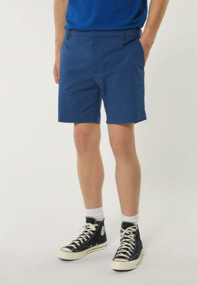 Shorts di cotone blu con un davanti piatto, tasche laterali e una superficie liscia, abbinati a sneakers alte nere e calzini bianchi.