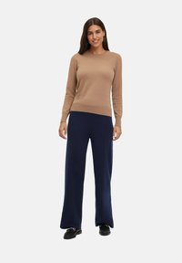 Beige gebreide trui met geribbelde manchetten, gecombineerd met marineblauwe wijde broek. Zwarte schoenen maken de outfit compleet tegen een effen witte achtergrond.