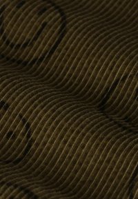 Tissu en velours côtelé brun avec des côtes verticales prononcées et un motif de visages souriants subtil, présentant une texture douce et des tons atténués.