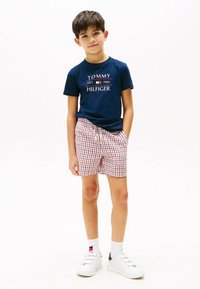 Marineblauwe katoenen T-shirt met zwart en rood 'TOMMY HILFIGER' logo, gecombineerd met rood en wit geruite shorts en witte sneakers.