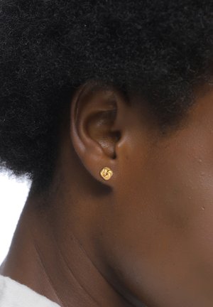 Boucle d'oreille en or avec un design en nœud torsadé, mise en valeur sur une peau sombre et des cheveux noirs bouclés. Fini en métal lisse, forme circulaire petite.