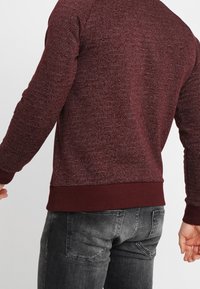 Bordeauxrotes Sweatshirt mit strukturiertem Strickmuster, gerippten Bündchen und Saum. Wird über dunkelgrauen Jeans getragen und verfügt über einen rund ausgeschnittenen Halsausschnitt.