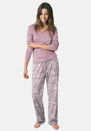 Kobieta uśmiechająca się i patrząca w dół, ubrana w długi rękaw lawendowej bluzki i luźne, kwieciste spodnie od piżamy w kolorze lawendy, boso.