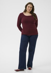 Bordeaux gekleurde ribbeltrui met lange mouwen en een gegolfde halslijn, gecombineerd met donkerblauwe uitlopende jeans en bordeauxkleurige ballerina's, tegen een effen achtergrond.