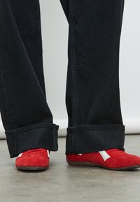 Pantaloni di denim nero con ampi risvolti abbinati a scarpe in suede rosso con dettagli bianchi. I pantaloni hanno una texture morbida e una vestibilità dritta.