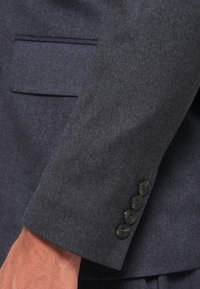Marine blazer med teksturert stoff, en enkelt brystlomme og fire svarte knapper på mansjetten for lukking.