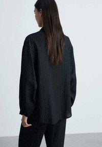 Polo a maniche lunghe nera con strisce verticali bianche, vestibilità rilassata e orlo leggermente arrotondato. Il tessuto sembra morbido e strutturato.