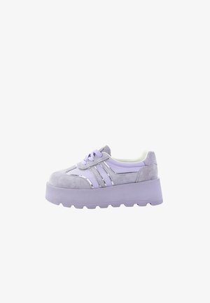 Gruba platforma sneakersów w jasnym fiolecie z zamszowymi i błyszczącymi paskami, wiązaniem na sznurówki oraz grubą podeszwą na białym tle.