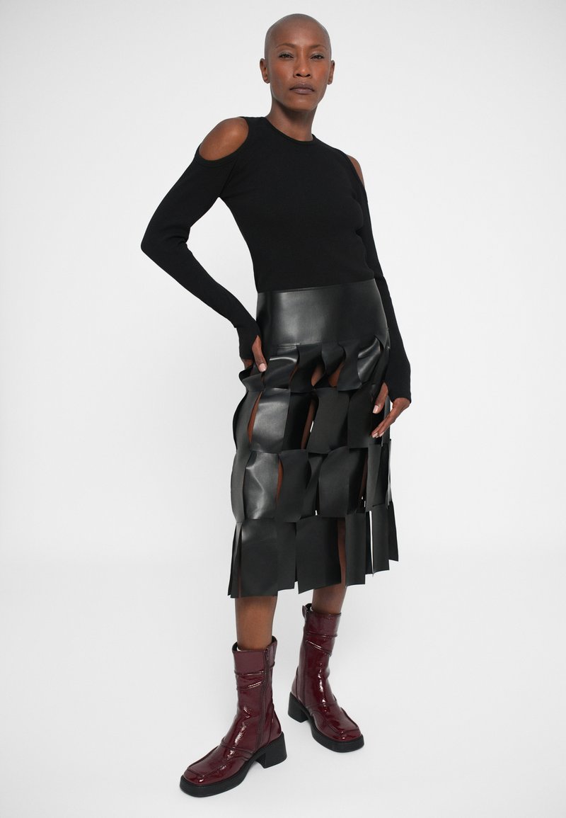 Vrouw in een zwarte cold-shoulder top, zwarte gestructureerde uitsnijrok en glanzende donkerrode laarzen tot halverwege de kuit, die zelfverzekerd tegen een effen achtergrond staat.