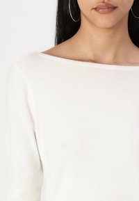 Long-sleeve, off-white top in a soft, smooth fabric with a wide neckline. Features a subtle texture and minimal design. Large hoop earrings visible.